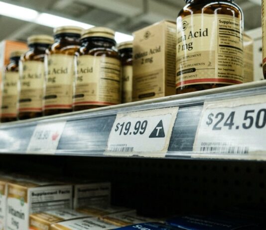 The T Is Back: What Loblaw’s Tariff Labels and Ottawa’s Counter-Tariff List Mean for Natural Health Grocery shelf label showing a triangle T tariff symbol beside a maple leaf Canadian product tag, supplement bottles above